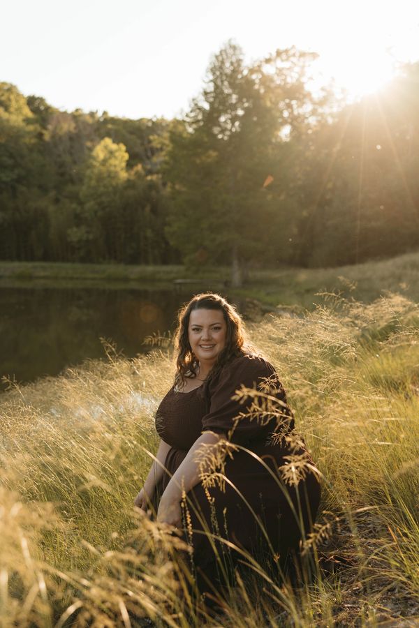 Woman sitting in a sunlit grassy field near a pond.