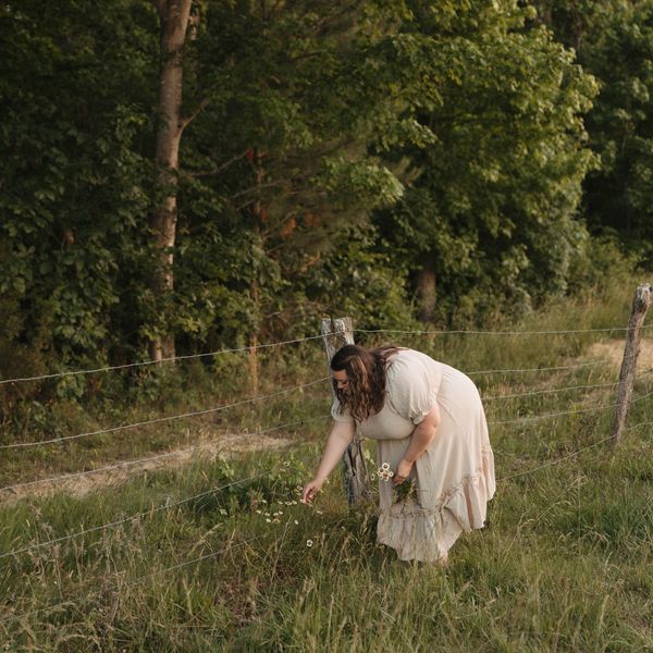 Woman in white dress picking flowers by fence.