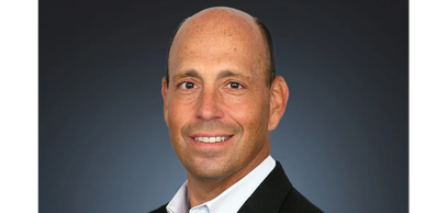 Professional headshot of a smiling man in a suit against a dark background.