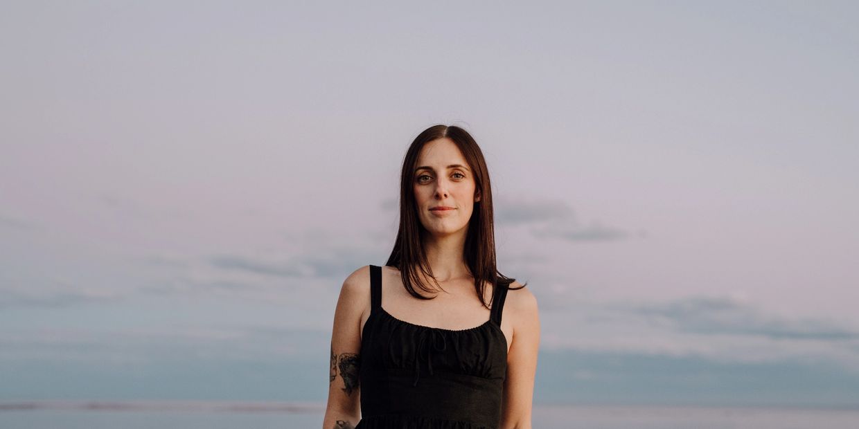 Young woman in a black dress standing against a serene pastel sky.