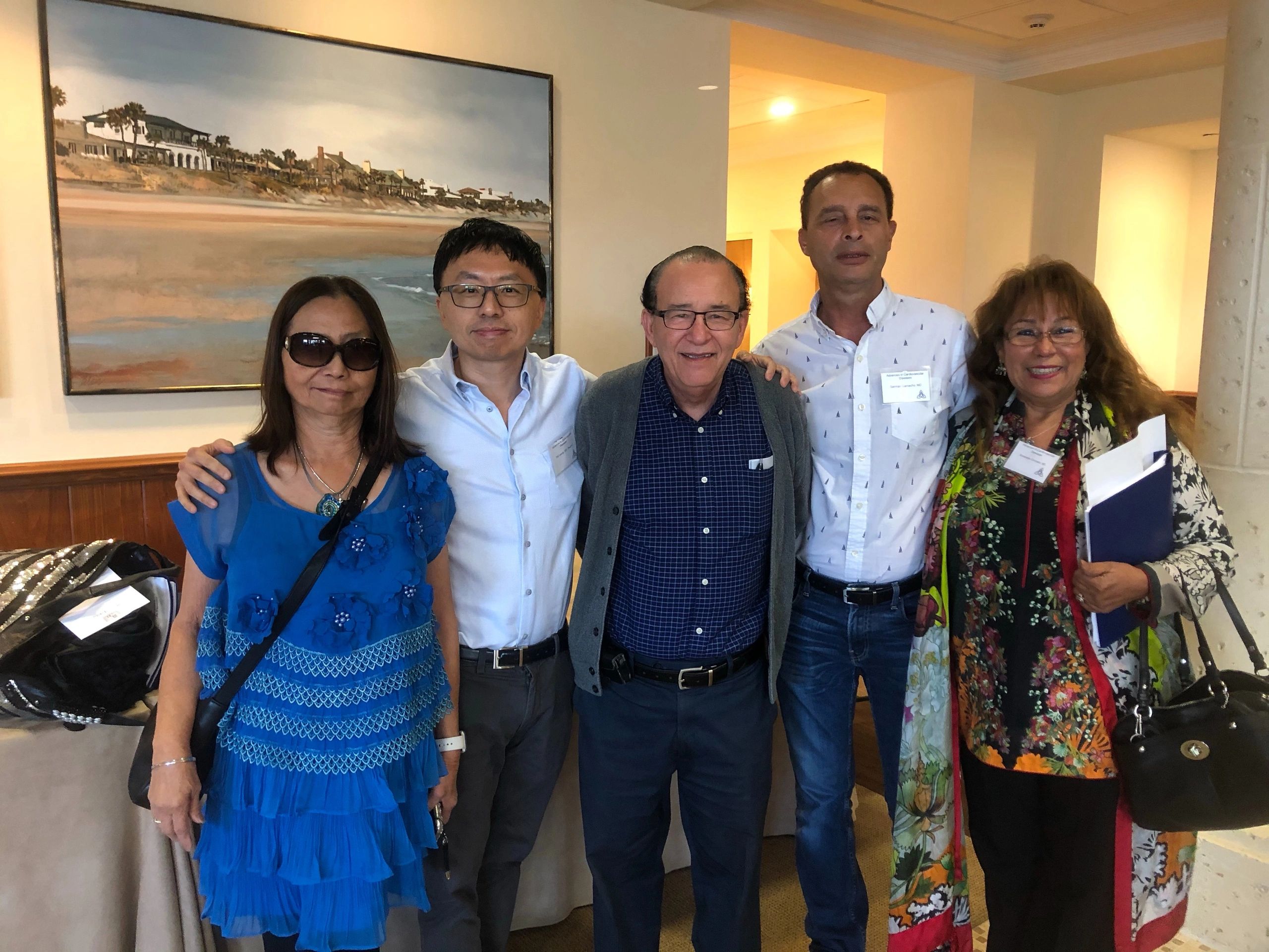 Five people smiling together indoors with a coastal painting in the background.