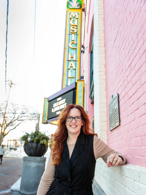 A smiling woman with red hair stands by a pink brick wall near a Music Hall sign.
