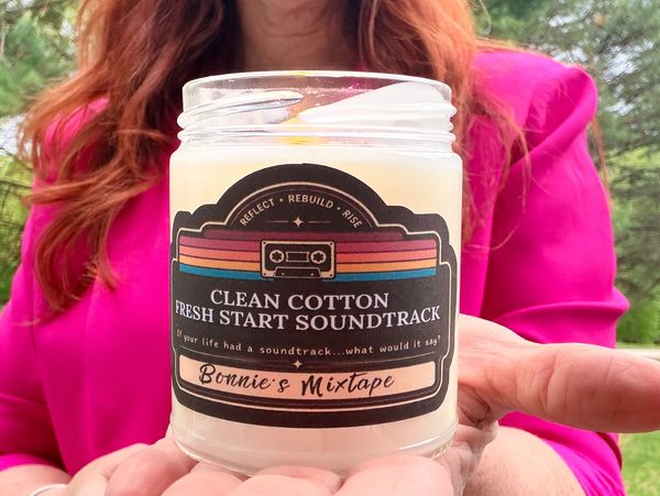 Person holding a scented candle labeled 'Clean Cotton Fresh Start Soundtrack'.
