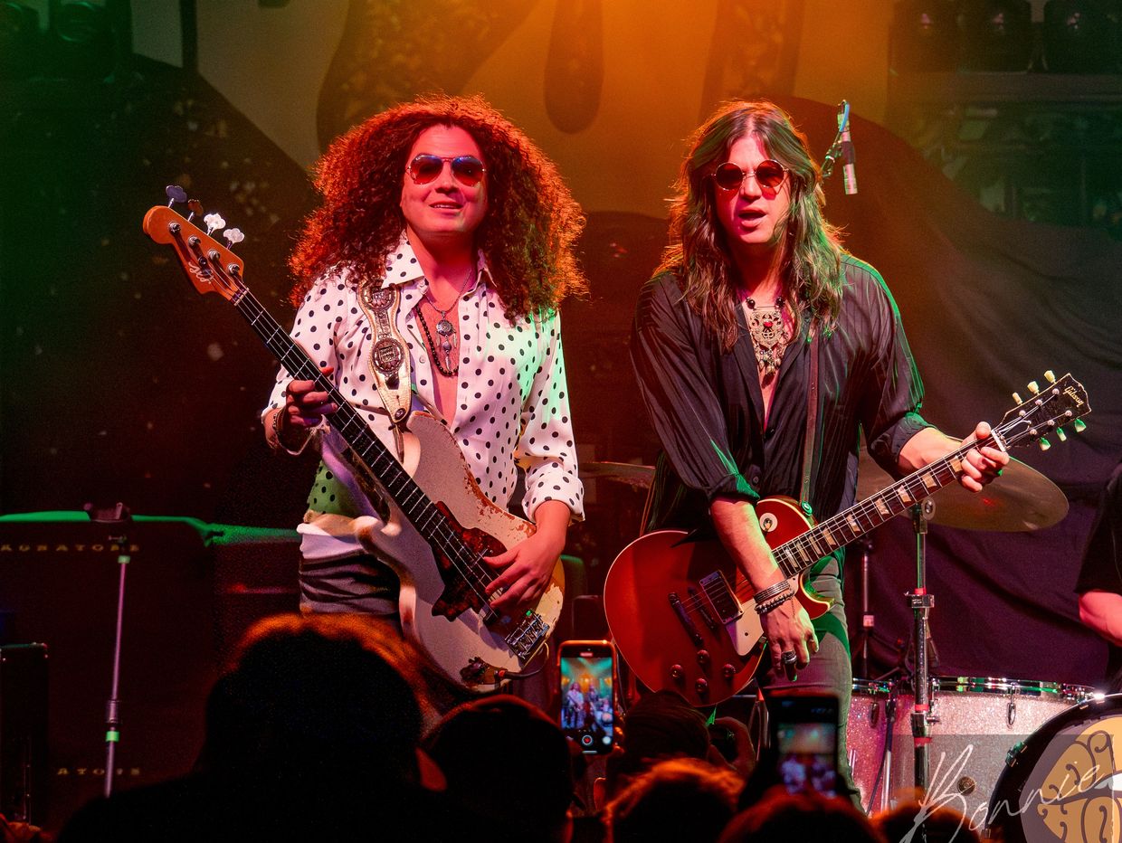 Two musicians with long hair and sunglasses playing guitars on stage.
