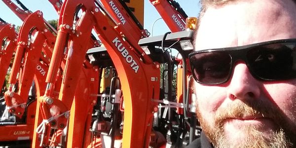 Man wearing sunglasses taking a selfie in front of orange Kubota excavators.