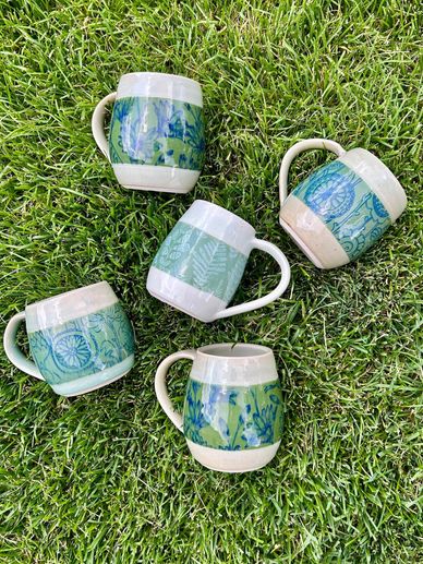 Five ceramic mugs with green and blue patterns arranged on grass.