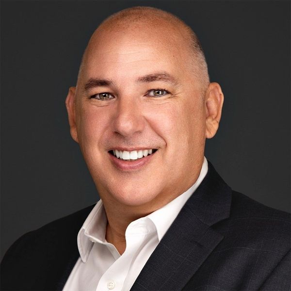 Smiling bald man in a suit jacket and white shirt against a dark background.