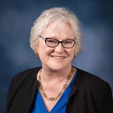 Smiling elderly woman with glasses in a blue top and black jacket.