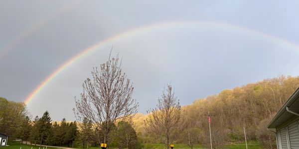 A vibrant double rainbow arches over a peaceful rural landscape with trees and a house.