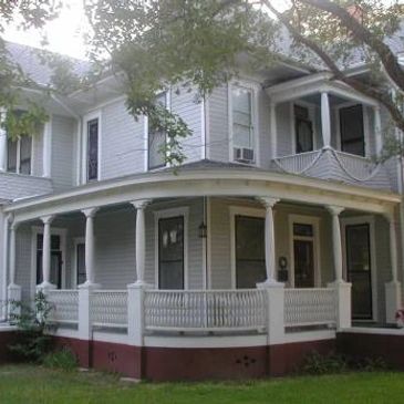 A large white two-story house with a wrap-around porch and mature trees.