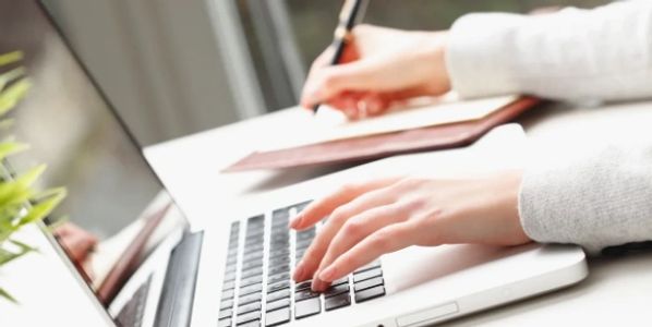 Person typing on a laptop while taking notes with a pen.