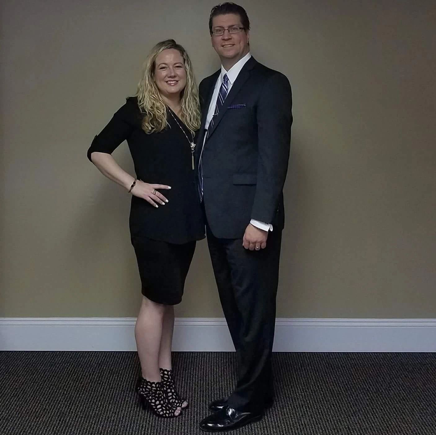 Couple smiling, dressed formally, standing against a beige wall.