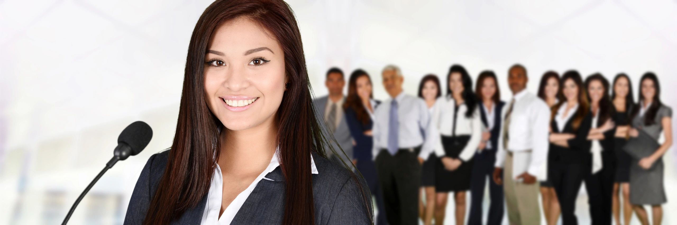 Young professional woman speaking confidently at a microphone with a business team behind her.