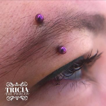 Close-up of eyebrow piercings with purple jewelry and eye makeup.