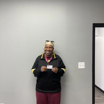Smiling person holding an ID card against a plain gray wall.