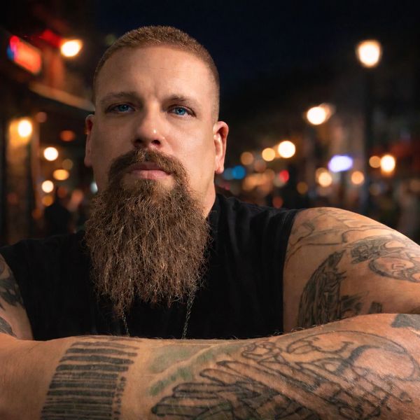 Bearded man with tattoos posing confidently at night with blurred city lights.