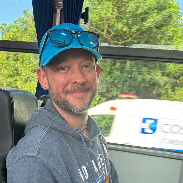 Man wearing a blue cap and sunglasses smiles inside a bus.