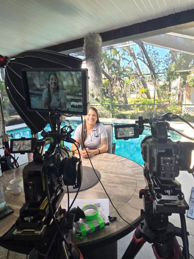 A woman is filmed during an outdoor interview by a pool with multiple cameras and a boom mic.