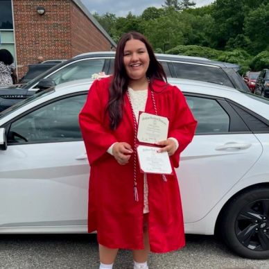 Young woman in red graduation gown holding diploma by a white car.