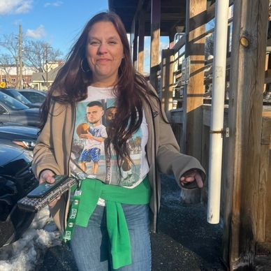Woman standing outside, smiling, wearing a graphic t-shirt and green sweater tied around waist.