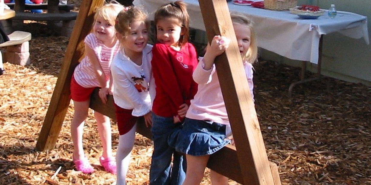 Four young girls playing together on a wooden swing set outdoors.