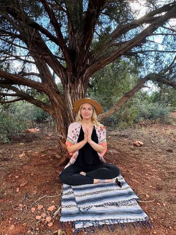 Woman meditating under a large tree on a patterned blanket outdoors.
