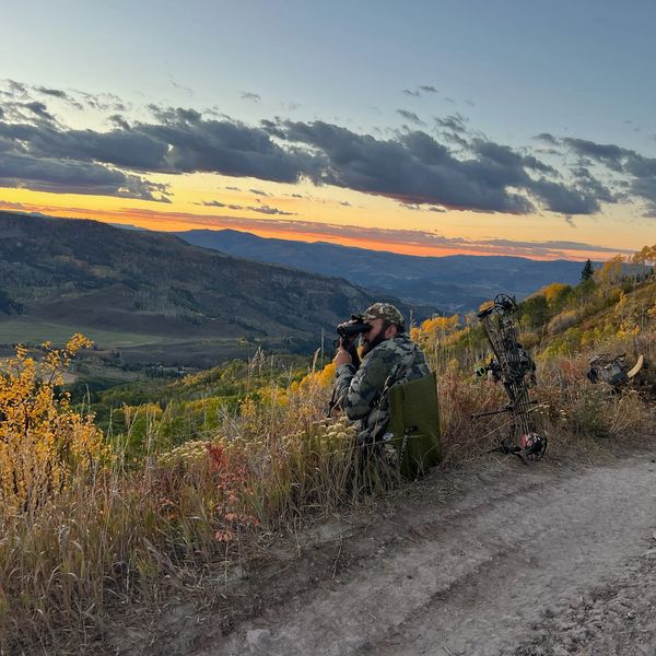 Hunter with binoculars overlooking a mountainous landscape at sunset.