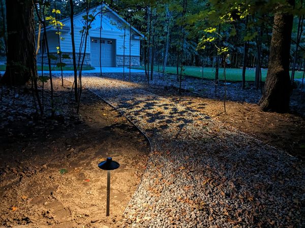 A lit pathway with gravel leading to a garage surrounded by trees at dusk.