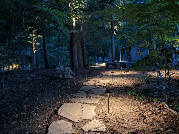 A stone path lit by garden lights leads to a cabin in a wooded area at dusk.