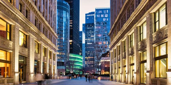 Urban street scene at dusk with illuminated buildings and people walking.
