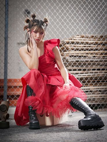 Young woman in a red dress and black boots sitting against a chain-link fence.
