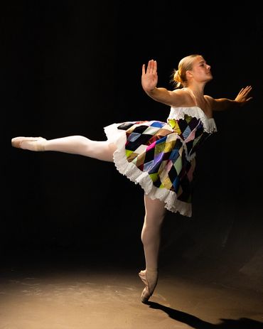 Ballet dancer performing an arabesque in colorful costume against a dark background.