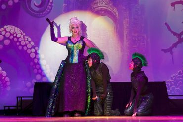 A woman in a glamorous costume performs on stage with two performers in dark, scaled outfits.