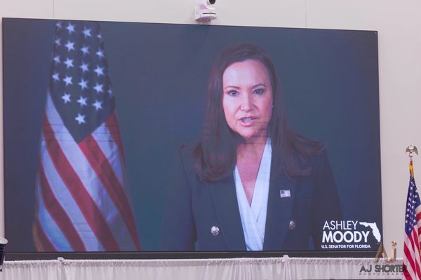 Ashley Moody addressing an audience through a screen with U.S. flags.