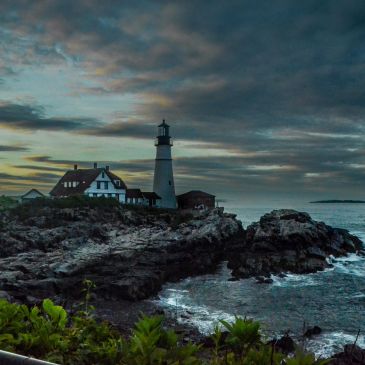 Lighthouse at rocky coastline during cloudy sunset.