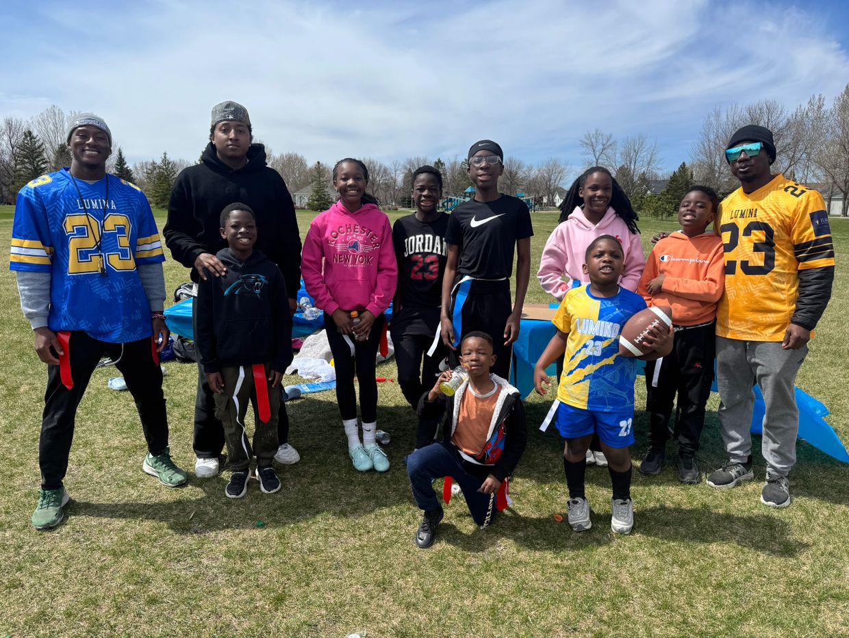 Group of kids and adults outdoors, some wearing sports jerseys and holding a football.