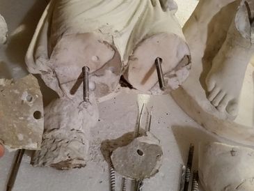 Hand holding a broken plaster piece near a damaged plaster statue with metal rods.