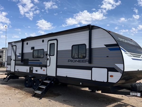 A modern Pioneer travel trailer parked on a dirt lot under a blue sky.