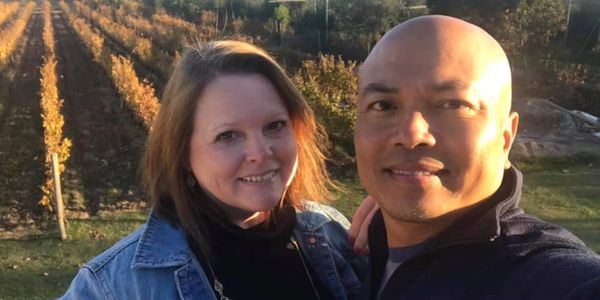 A couple smiling outdoors with vineyard rows in the background during sunset.