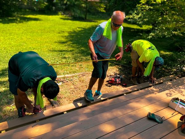 Three men measuring and assembling wooden planks outdoors under sunlight.