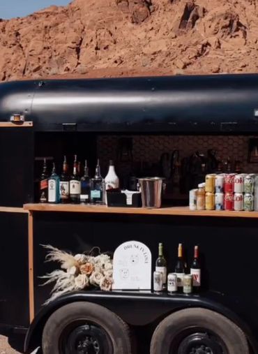 Outdoor mobile bar setup with beverages and floral decoration against rocky terrain.