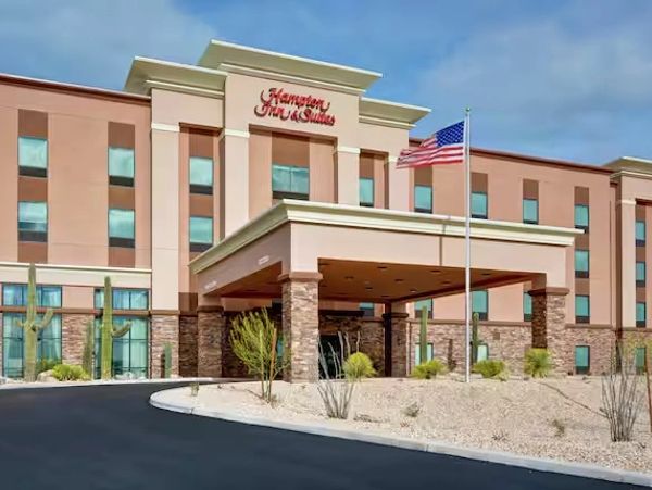 Exterior of a Hampton Inn &amp; Suites hotel with desert landscaping and American flag.
