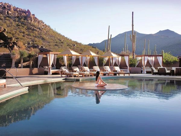 A woman relaxing by a serene pool with desert mountains and cacti in the background.