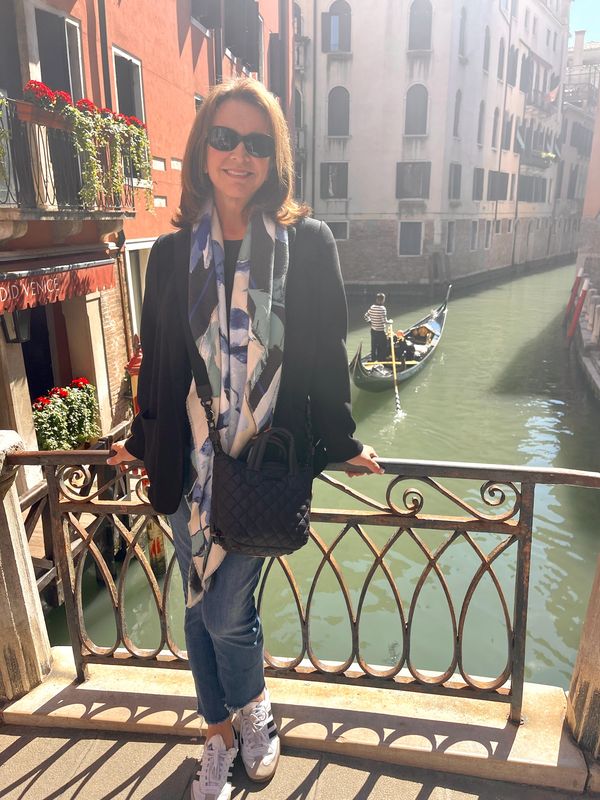 A woman stands on a Venetian bridge with a gondola in the canal behind her.