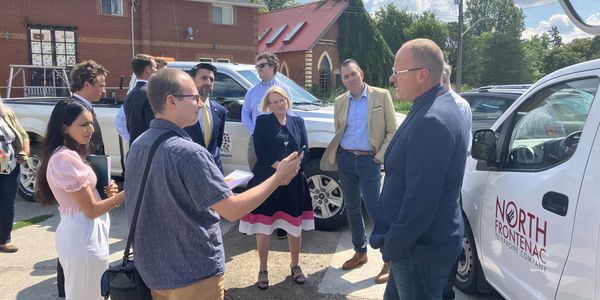 Group of people in business attire conversing outside near vehicles on a sunny day.