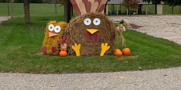 Festive hay sculptures of a turkey, chick, and scarecrow with pumpkins on a grassy lawn.