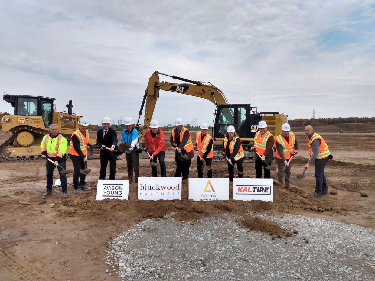 Group of people in hard hats and safety vests at a groundbreaking ceremony with construction equipment.