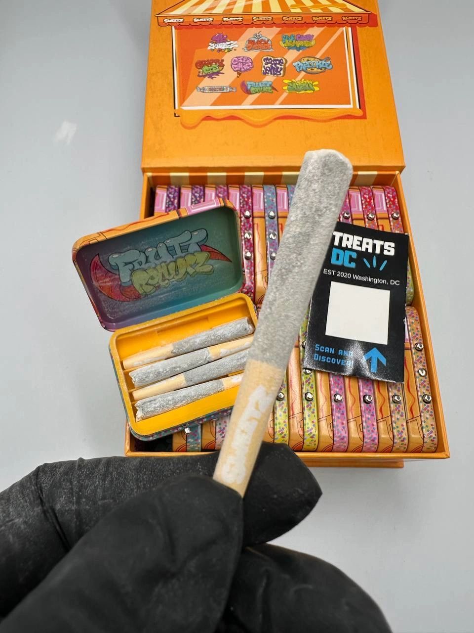 A gloved hand holding a pre-rolled joint with a colorful box of more joints in the background.
