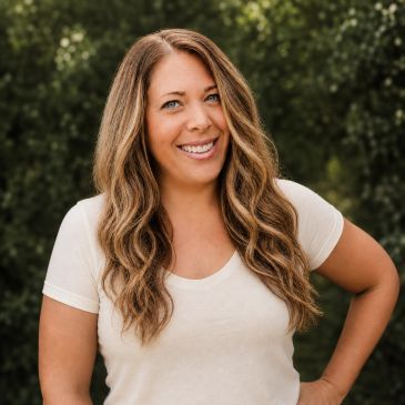 Smiling woman with long wavy hair wearing a white t-shirt outdoors.