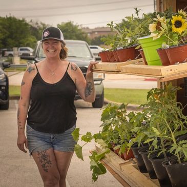 Smiling woman standing beside a shelf of potted plants outdoors.
